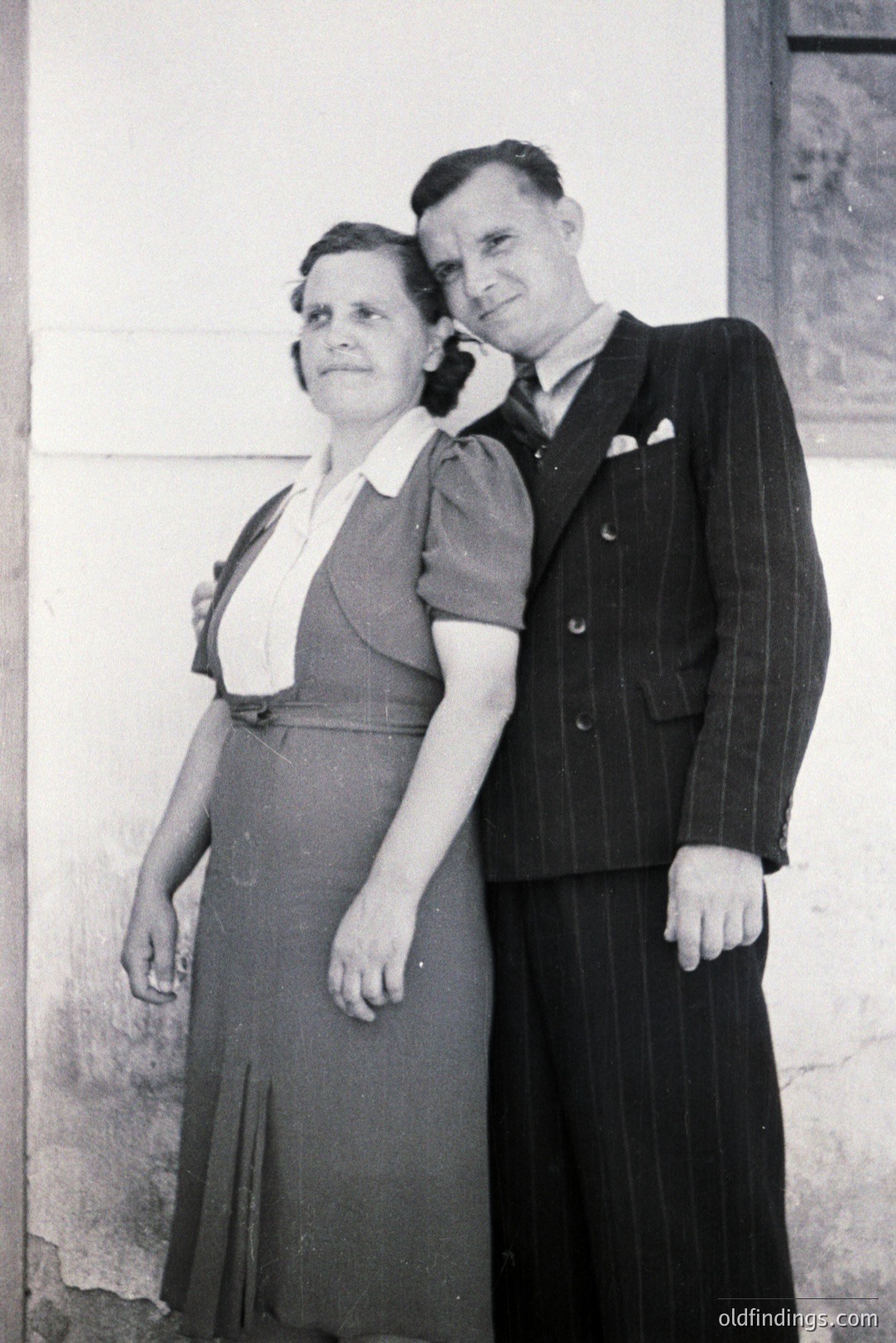 A formally attired couple poses outside a stucco building. The woman wears a modest, knee-length dress with a contrasting collar, while the man is in a pinstriped suit. Likely a portrait from the 1940s or 50s. Offers archival value related to mid-century fashion and family photography.