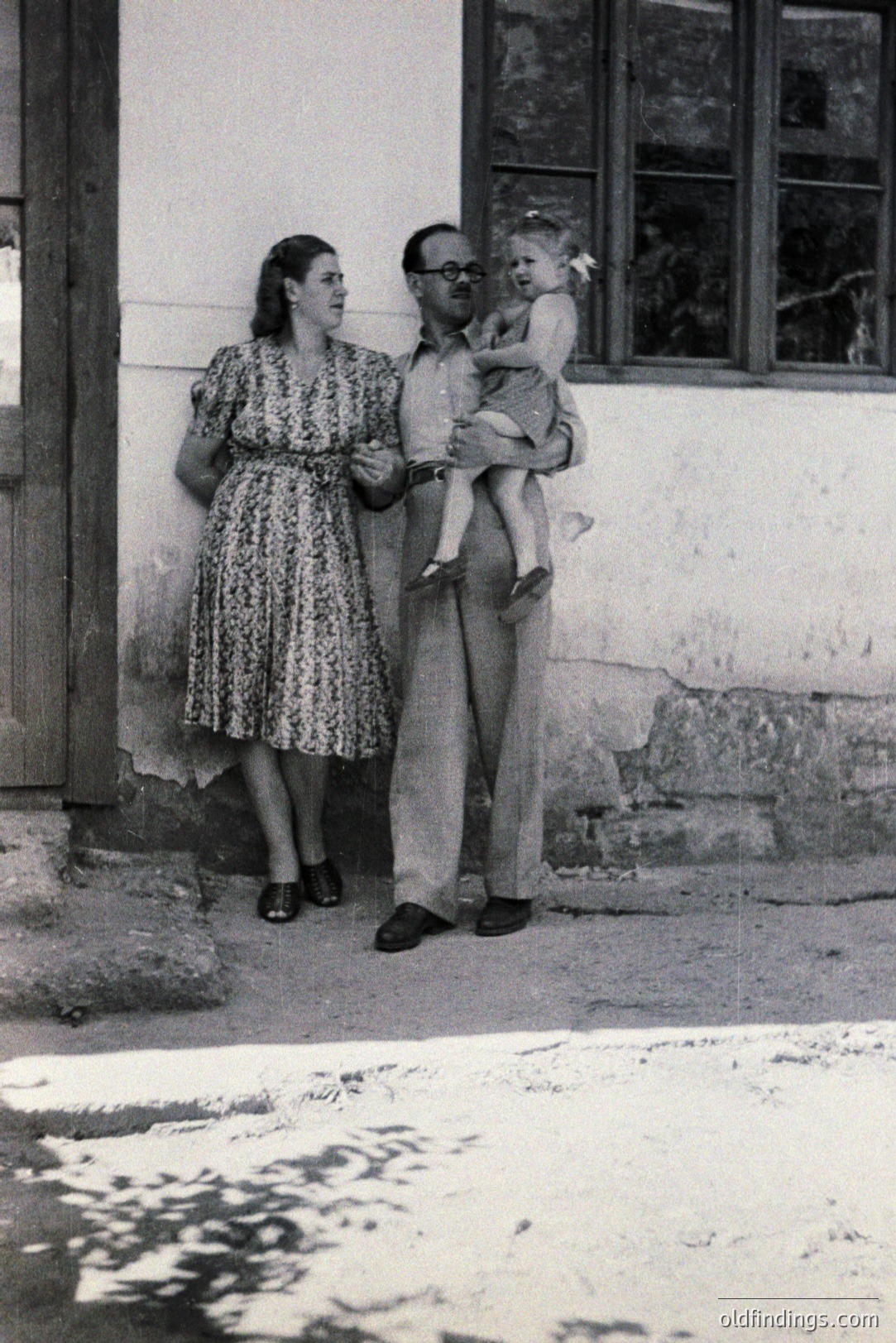 A family portrait, likely 1950s or 60s. A man in a short-sleeved shirt and trousers holds a toddler; a woman stands beside them in a patterned dress. Exterior scene with rustic wall and window visible. Suggests a rural or vacation setting. A moment of family life captured in time.