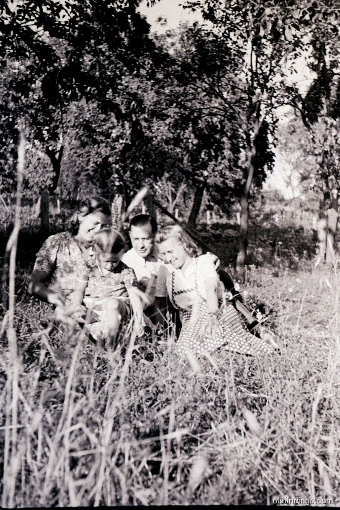 A family portrait, likely mid-1940s, shows a woman holding a young child, alongside a teenage boy. All three are nestled within tall grasses and foliage. Casual attire suggests a relaxed, outdoor setting. Simple composition, characteristic of family snapshots. Potential value for genealogy or historical context.