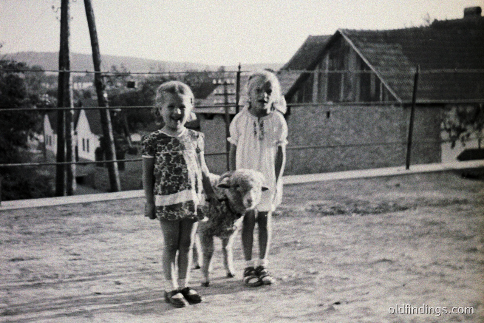 Two young girls, one in a floral dress, the other in a plain dress, stand alongside a small lamb. A rustic wooden fence and traditional buildings form the backdrop. Likely a rural setting, potentially Eastern Europe. Appears to be a candid family snapshot, c. 1950s-1970s. Rural childhood memories.