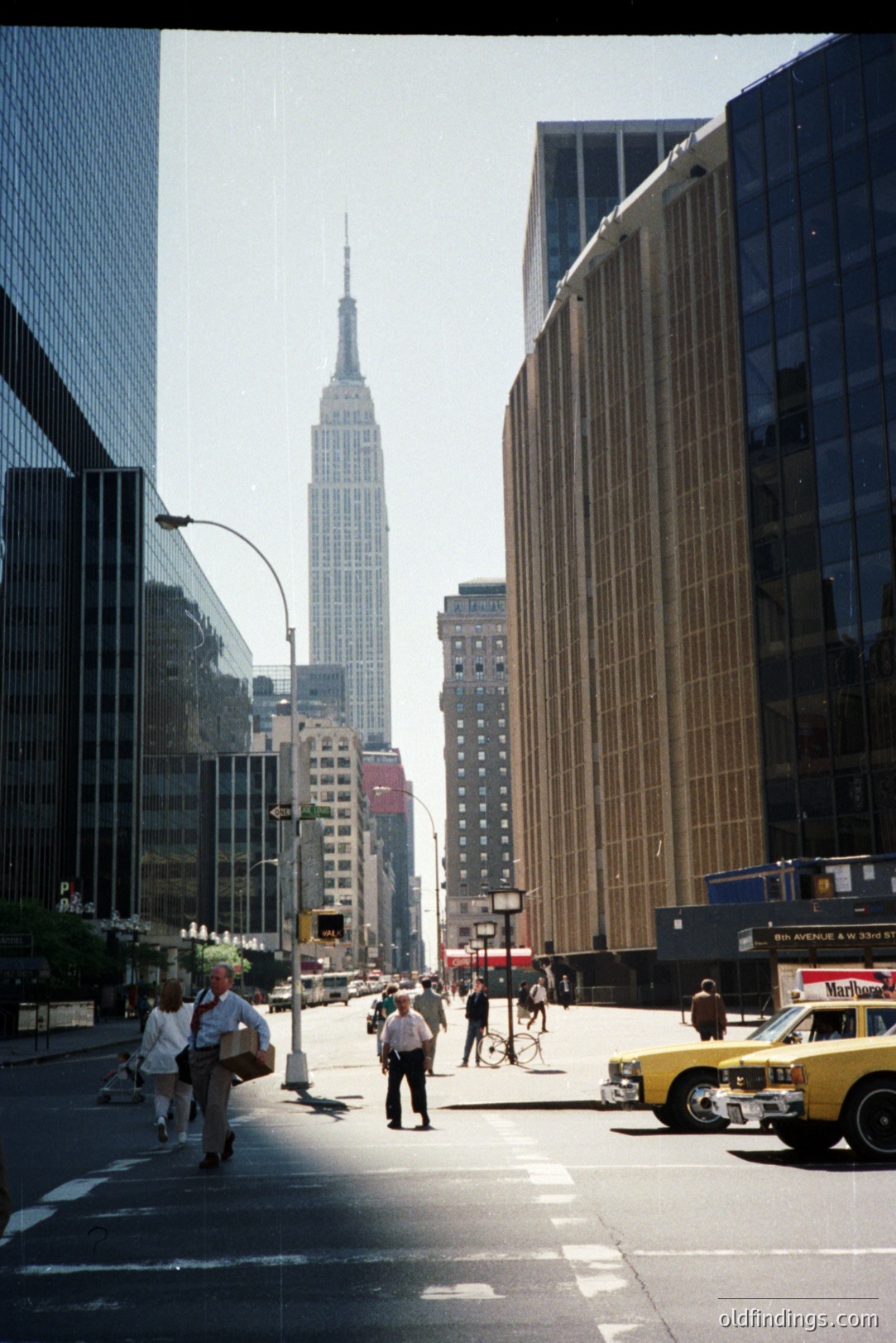 A view down a New York City street toward the Empire State Building, framed by distinctive mid-century architecture. A yellow taxi cab and pedestrians populate the foreground. Likely 1960s-1970s, judging from the vehicles and style. Commercial potential for architectural reference & NYC cityscape imagery.