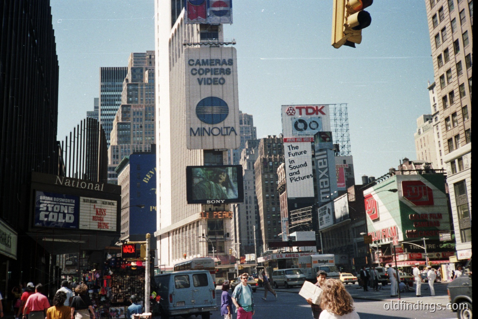 Times Square, NYC, showcasing vintage signage. The Minolta and TDK billboards are prominent, indicative of 1980s consumer tech. People & vehicles populate the street; a busy urban scene. A moment captured reflecting a bygone era of advertising.