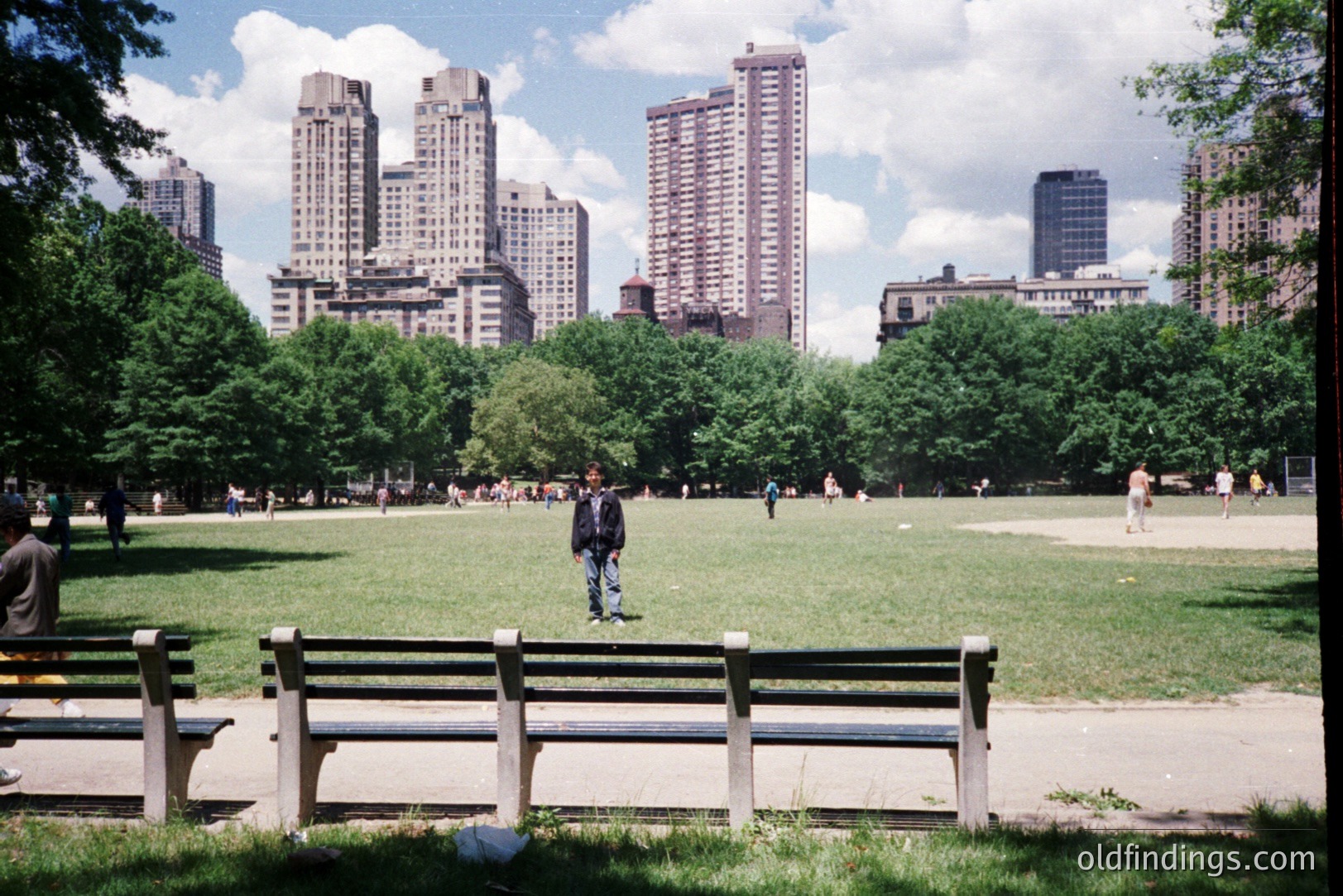 A vibrant, wide-angle view of a park with a man walking across the grassy expanse, framed by trees and a backdrop of dense urban high-rise buildings. Likely Central Park, New York City. The scene features a concrete pathway and bench in the foreground. Appears to be a candid shot, possibly 1980s or 90s film.