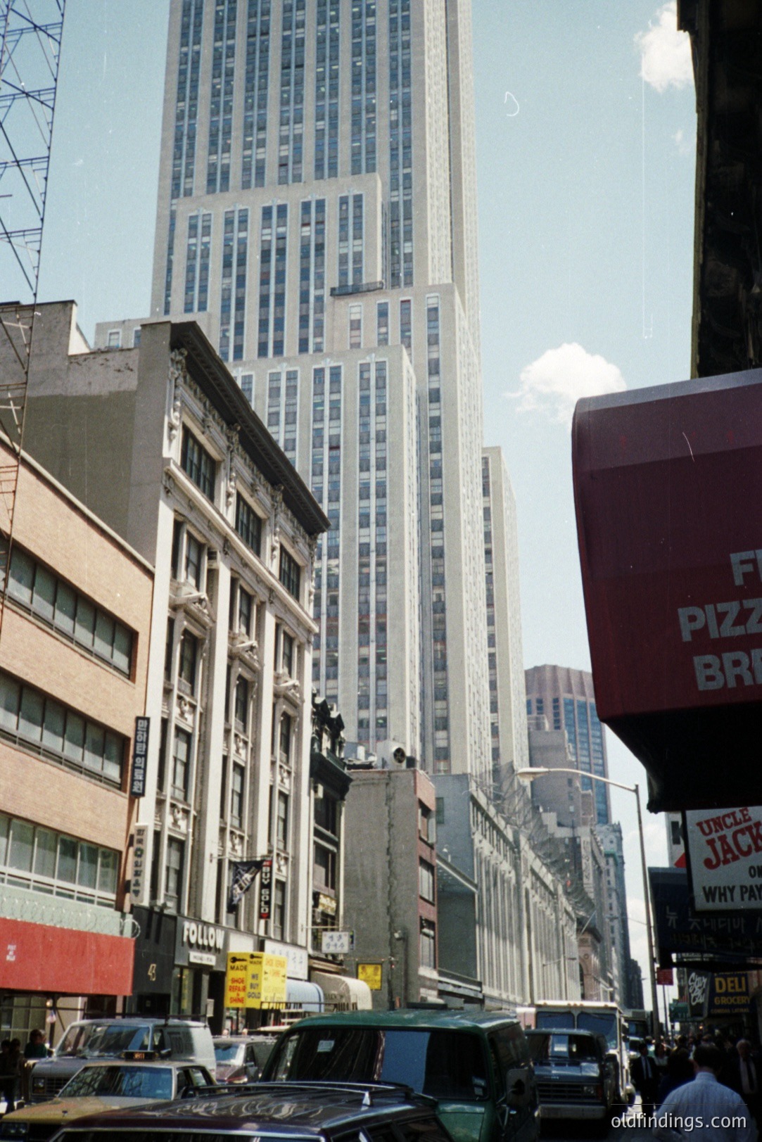 A street view in New York City, circa 1970s. Classic architecture lines the block, dwarfed by the Empire State Building rising prominently in the background. Vintage cars navigate the busy street, alongside storefront signage. A typical urban landscape.