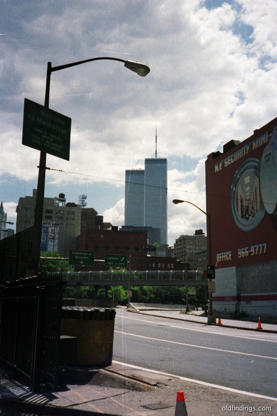 Overpass view showcases a "Welcome to Manhattan" sign and the iconic, then-standing, World Trade Center towers in the background. Visible signage indicates the FDR Drive. Commercial signage includes "NY SECURITY MINI-SAFE". Likely taken pre-9/11. Documenting urban infrastructure.