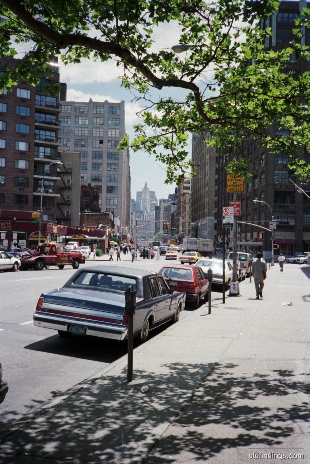 A 1970s-80s urban street scene in New York City. Classic cars line the curb; pedestrians populate the sidewalk and crosswalk. Tall apartment buildings dominate the background with the Empire State Building visible in the distance. Likely taken in the 1980s, showcasing a distinct mid-century aesthetic.