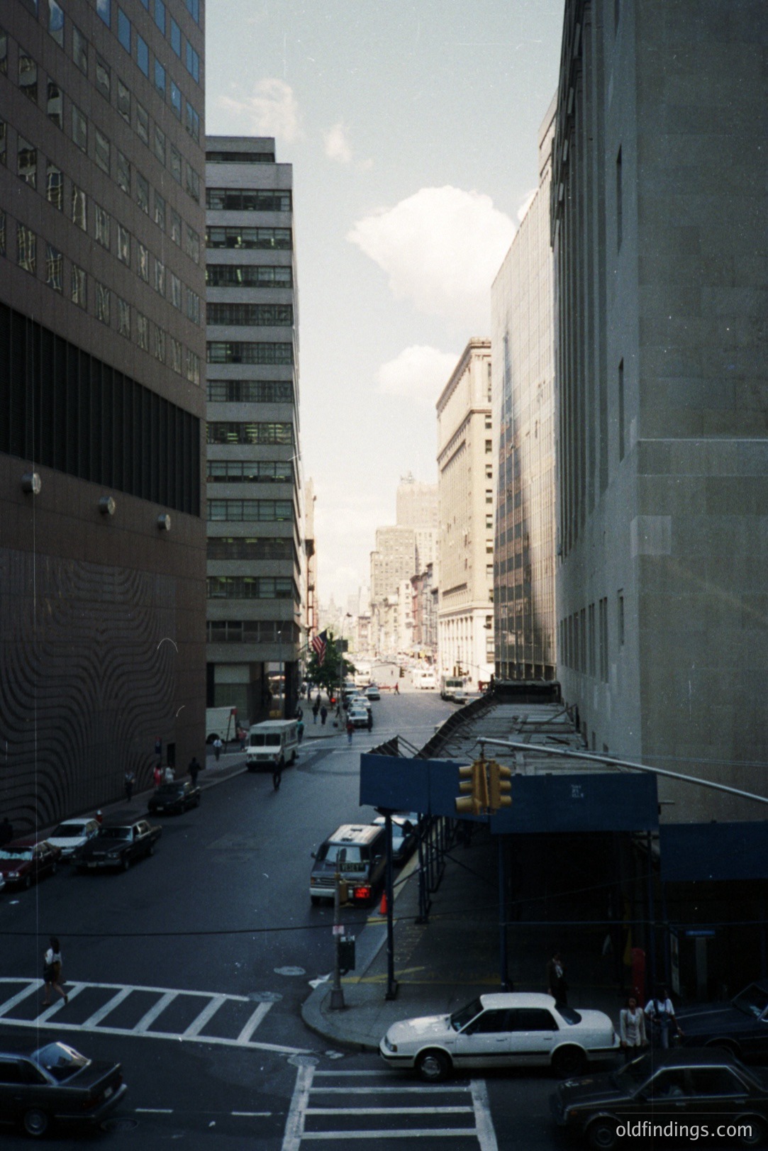 Looking down a city street framed by towering, modernist office buildings. Visible cars and pedestrian traffic suggest urban activity. Likely taken in the 1970s or 80s, judging by vehicle styles and architectural details. Potential commercial value for urban design references.