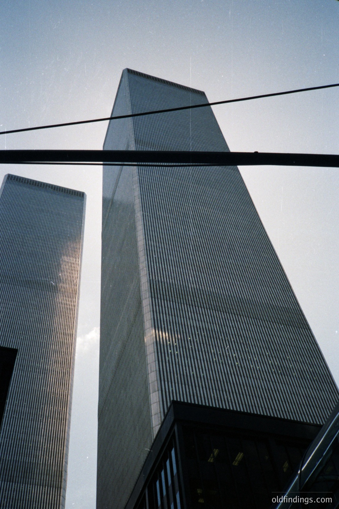Looking up at the iconic World Trade Center towers in New York City. A low-angle shot showcases the buildings' immense height & distinctive architecture. Visible power lines cross the frame. Likely taken in the 1970s-1990s. Significant historical & architectural value.