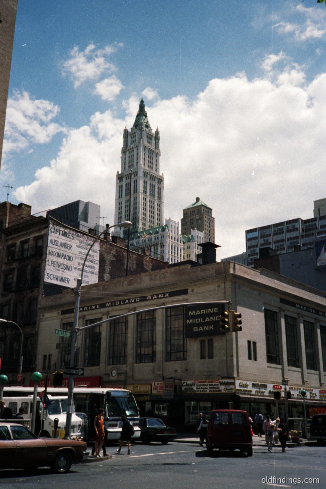 Classic 1970s street scene in New York City. A Marine Midland Bank building is prominent, dwarfed by a taller Art Deco skyscraper. Visible signage includes "Schirmer" and "Petrosino." Traffic and pedestrians populate the urban intersection. A vibrant, nostalgic snapshot of downtown architecture.