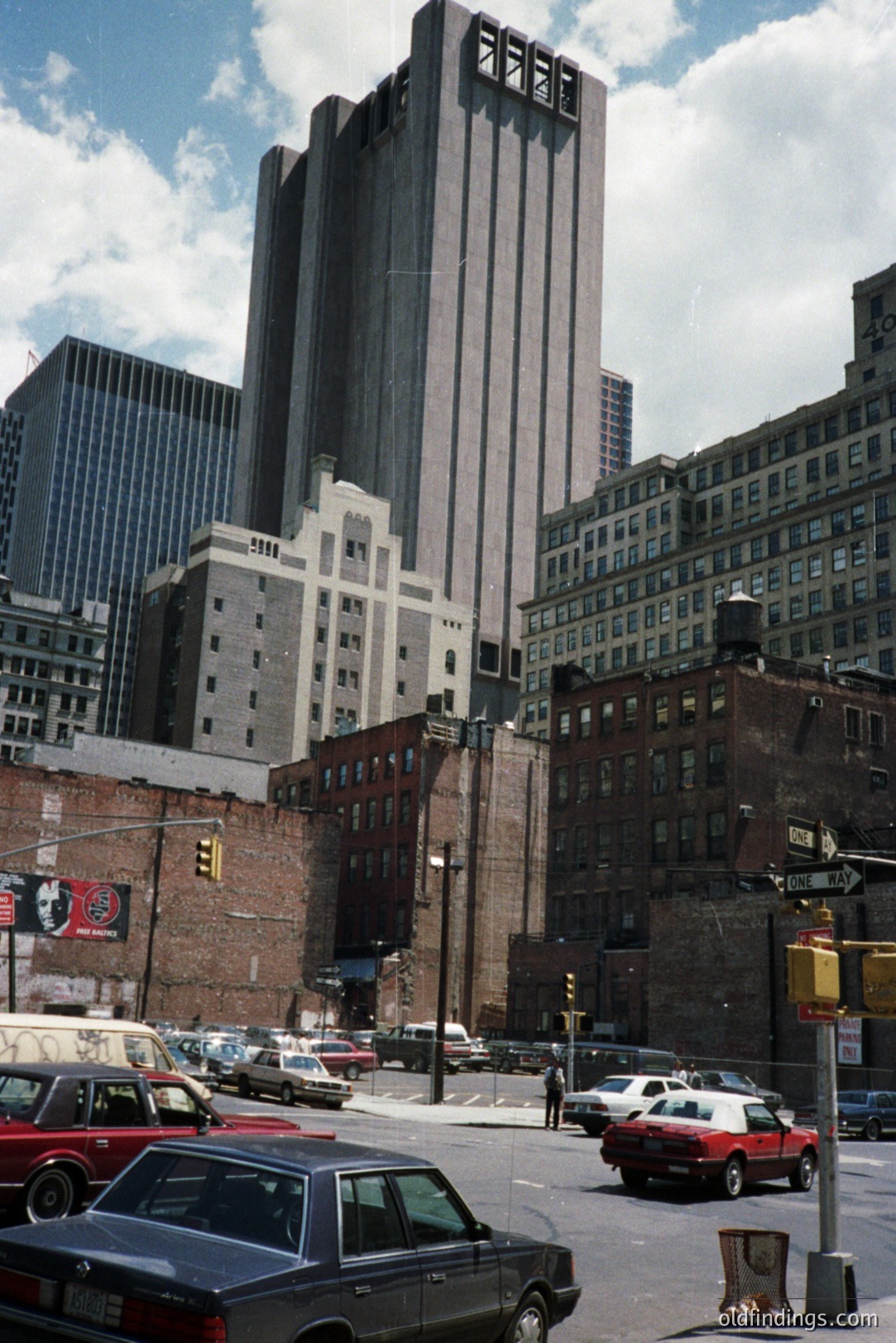 A view of a distinctive, tiered building in downtown New York City. Red brick buildings and a billboard flank the foreground. Traffic and pedestrians are visible on the street. Appears to be a 1970s or 80s photograph. Historic architecture and urban landscape.