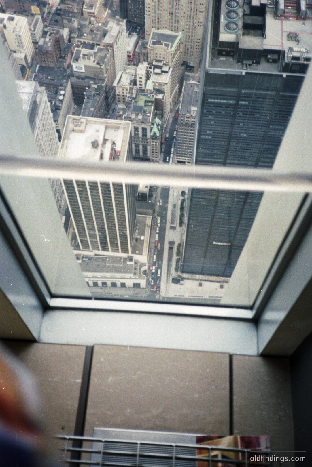 High-angle view through a window shows a cityscape with densely packed buildings and visible street traffic. The frame includes structural elements of the building and a portion of a person's arm, indicating a vantage point from a high floor. Likely New York City.
