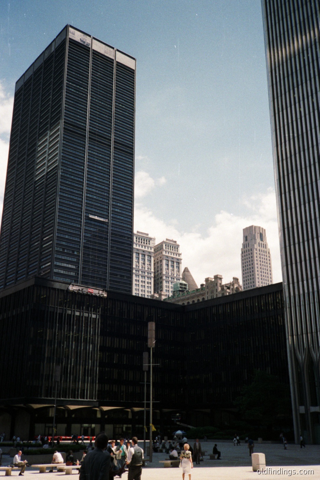 Striking modernist architecture frames a view of New York City skyscrapers. The foreground building exhibits a grid-like façade, typical of 1960s corporate design. Visible crowds populate the plaza. A classic example of urban design & architectural interplay.