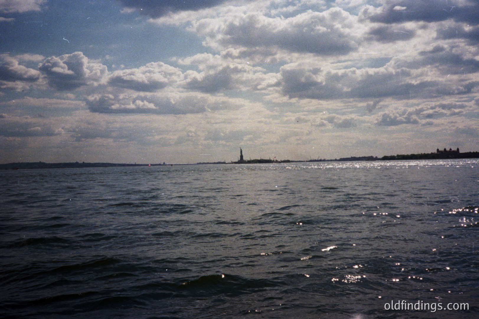 A long, wide shot captures the Statue of Liberty across a shimmering body of water, likely New York Harbor. The sky is overcast with dramatic cloud formations, reflecting on the choppy water's surface. The distant shoreline is visible, adding depth. Likely a tourist photo from the 1980s or 90s.