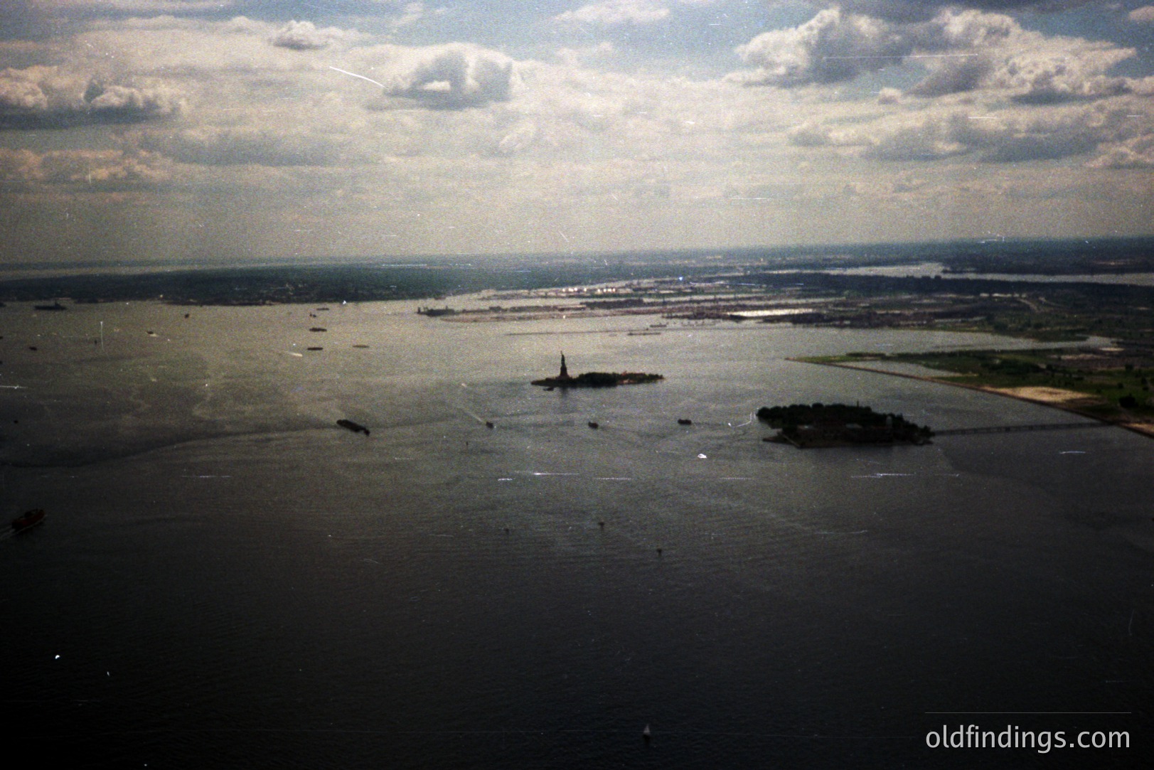 Aerial view of New York Harbor showcasing Liberty Island with the Statue of Liberty, Ellis Island, and extensive shipping traffic. The background shows a developed shoreline. Likely mid-to-late 20th century due to film quality and vessel types. A classic view of a major port.