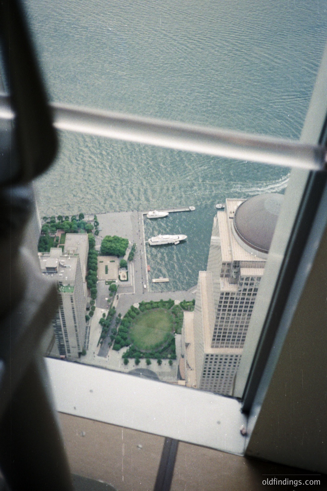 High-angle view through a window reveals a harbor scene featuring a modern building with a domed roof, likely a convention center. A luxury yacht is docked alongside a pier, framed by manicured lawns and waterfront landscaping. Appears to be an urban coastal setting.