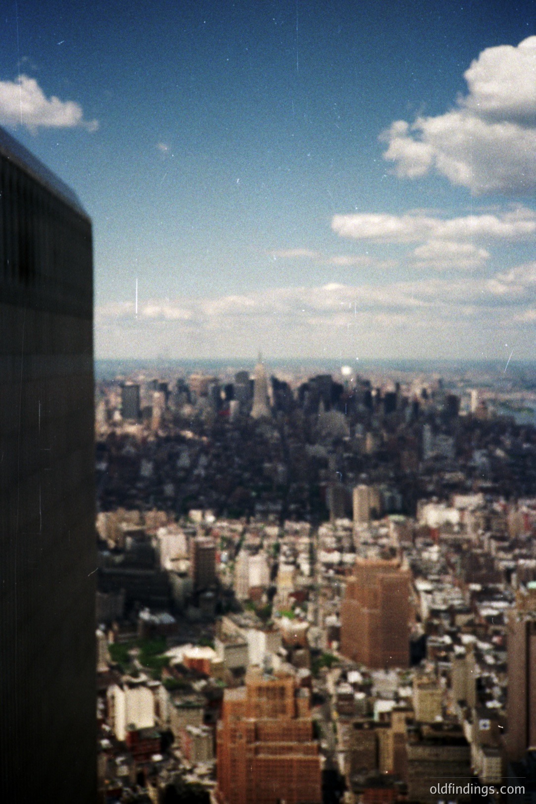 High-angle view of lower Manhattan, NY, featuring the Empire State Building & a partial view of the Statue of Liberty. Likely taken from a skyscraper, showcasing a densely-built urban landscape under a partly cloudy sky. Appears to be film photography, potentially 1970s-1980s. Architectural documentation/stock reference.