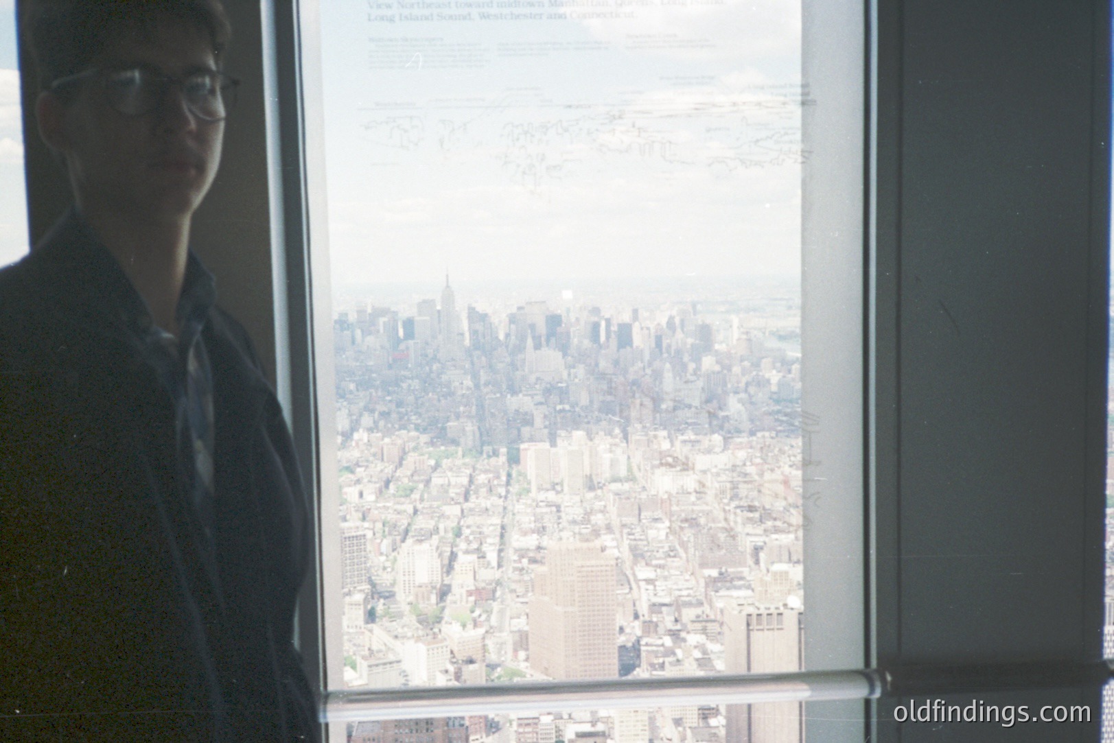 A person in a dark jacket & glasses stands near a large window overlooking a dense urban landscape, likely New York City. Visible buildings suggest a mid-20th century viewpoint, possibly from an observation tower. The image exhibits a slightly faded color palette, indicating potential age.