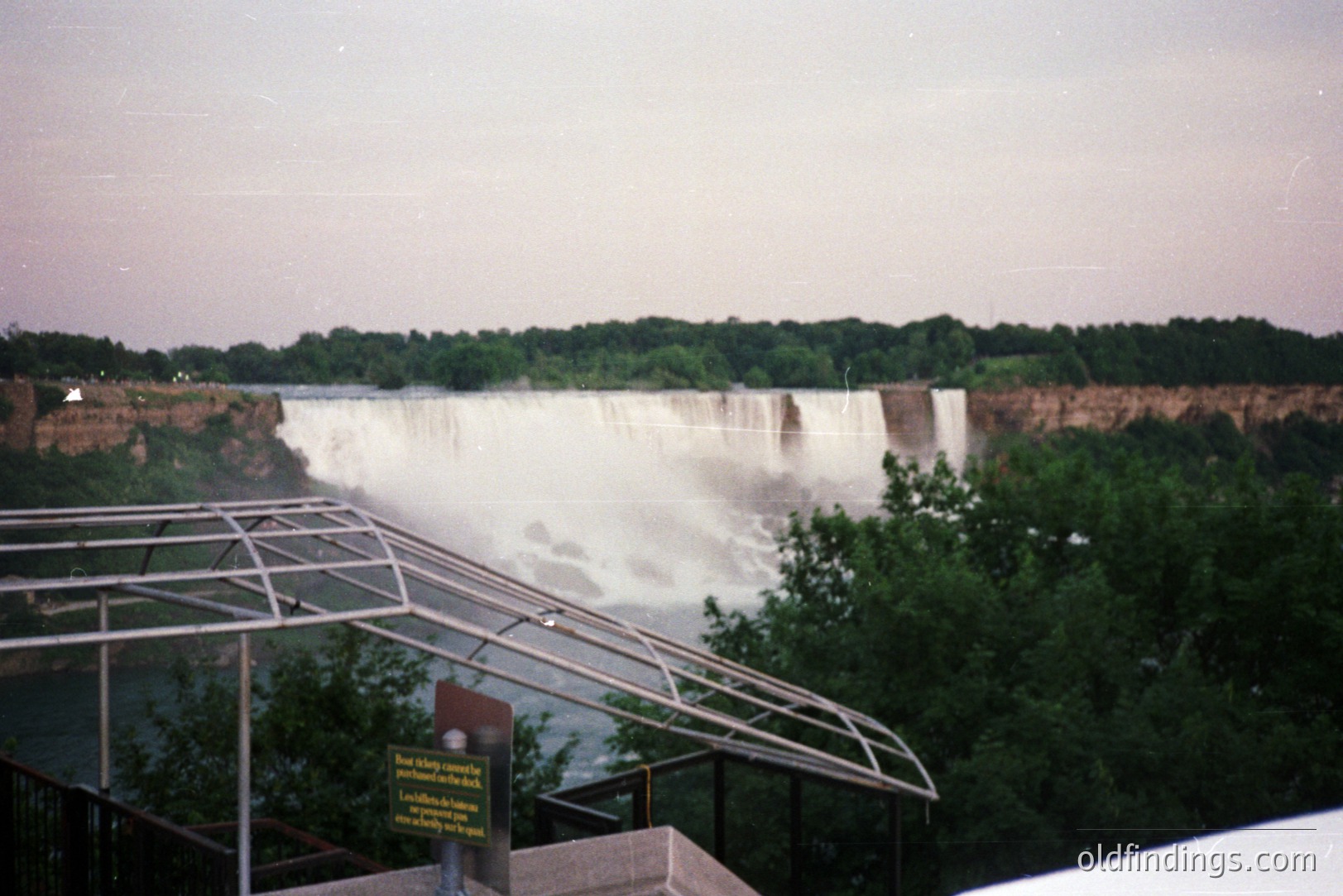 Spectacular view of Niagara Falls, partially obscured by mist. A viewing platform with a modern railing and informational signage is in the foreground. Lush greenery frames the falls. Likely a tourist photo, mid-late 1990s based on image quality.