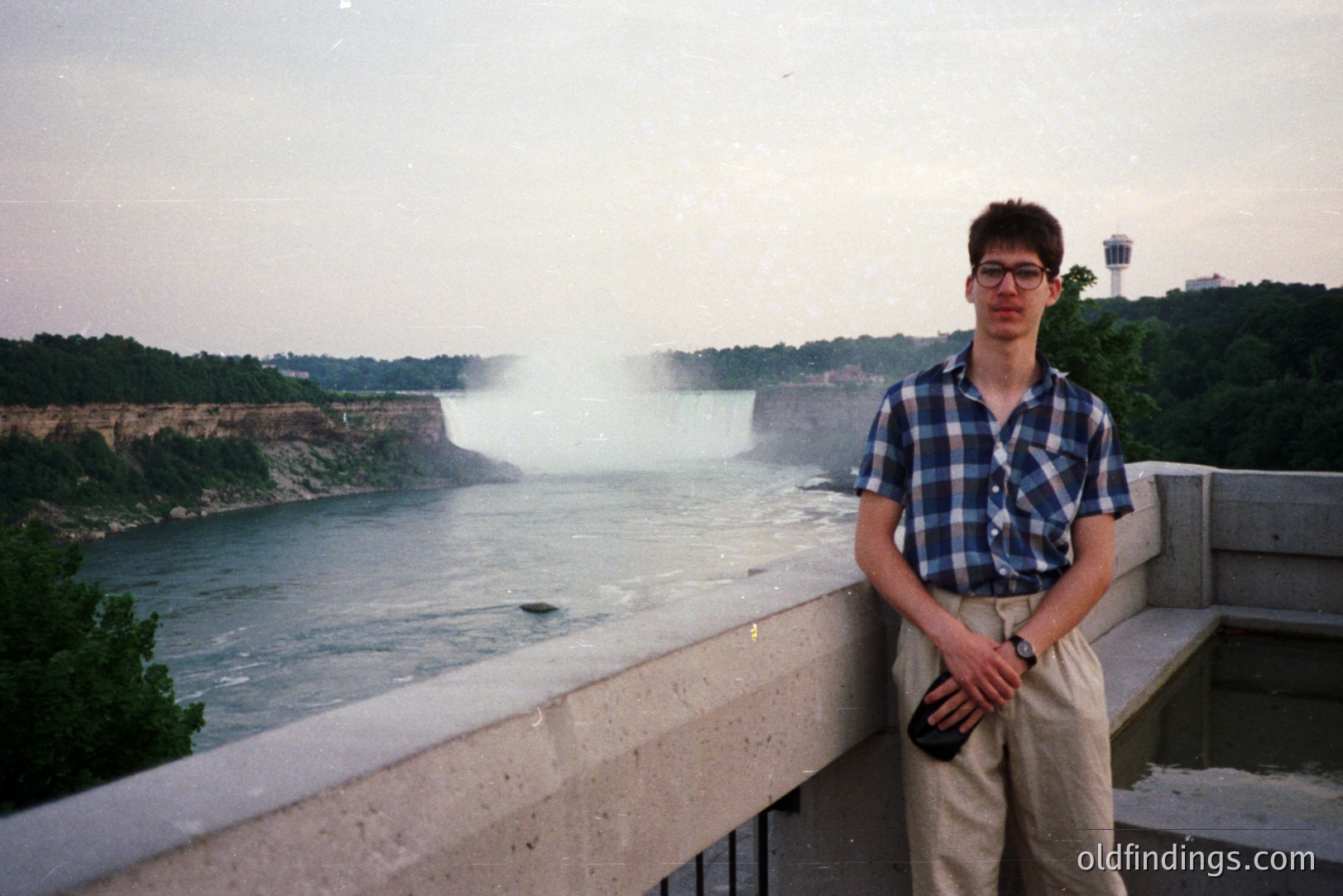 Young man stands before Niagara Falls. He wears a plaid shirt, khaki pants, and glasses. Concrete railings separate viewer from the falls, creating a misty effect. Likely a tourist photo, captures mid-1990s aesthetic. Potential stock photo reference for travel or portraiture.