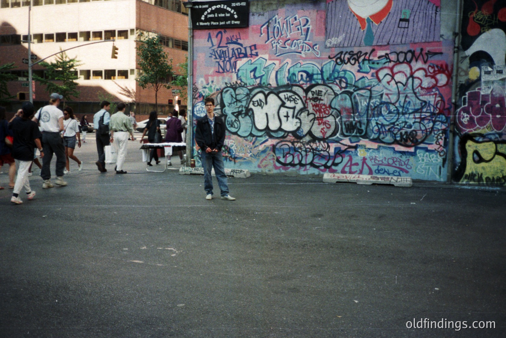 A young man stands center frame on a paved area with graffiti-covered wall backdrop. Several people walk beyond him. The artwork includes vibrant tags and cartoon imagery. Likely urban setting; appears to be a public space. Photographic style suggests 1990s. Commercial value: urban design, street style reference.