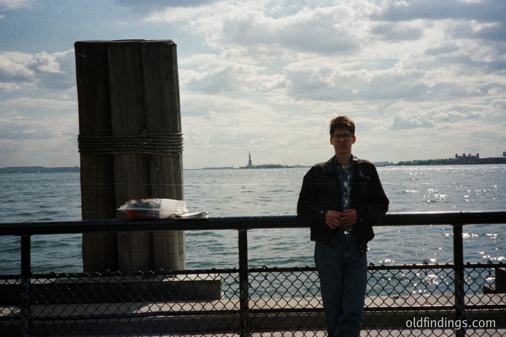 A young man stands near a pier, gazing toward the Statue of Liberty and the Manhattan skyline. He wears a dark blazer and jeans, a style typical of the 1990s. The scene features a weathered pier with a large wooden piling. Likely taken in New York City.