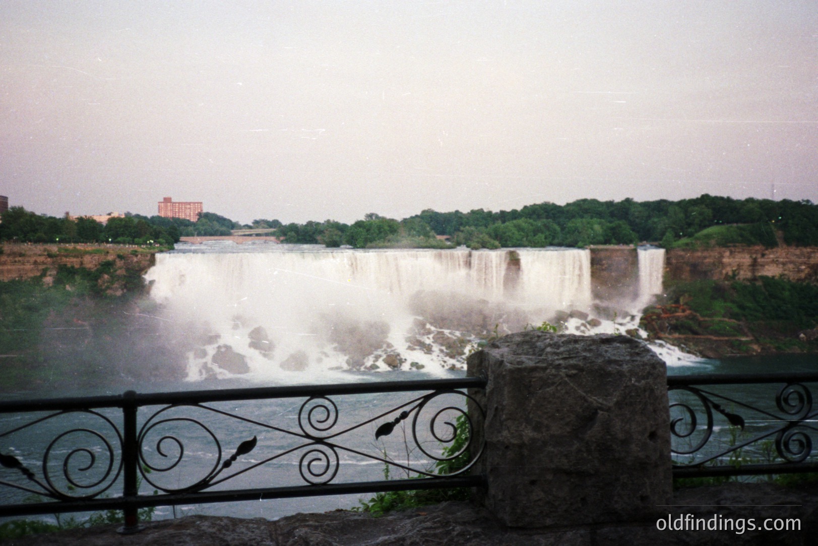 Dramatic view of Niagara Falls, showcasing the powerful cascade of water and misty spray. Lush green trees frame the falls, with buildings visible along the horizon. Ornamental railing and a stone block create a foreground detail. Likely 1970s or 80s based on film aesthetic.