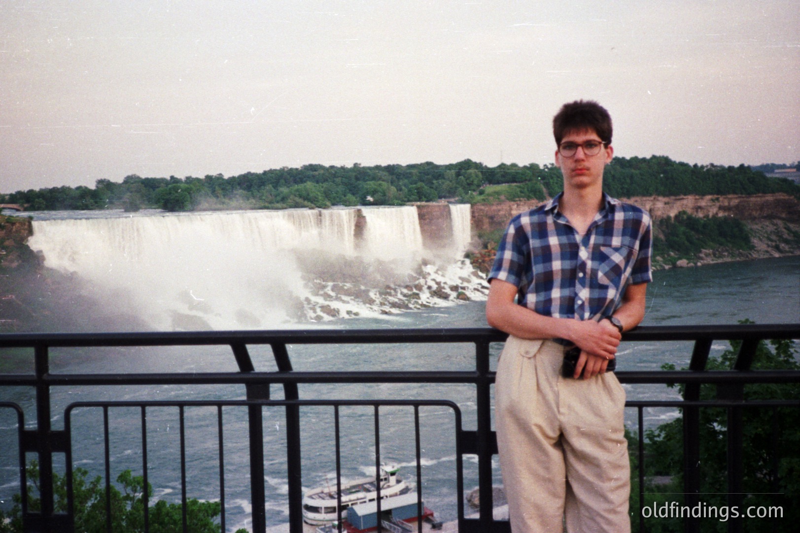 Young man in a plaid shirt and high-waisted trousers poses before Niagara Falls. A boat tour vessel is visible near the base of the falls. Likely a tourist photo from the 1980s. The image has a dated, slightly grainy aesthetic. A classic travel snapshot.