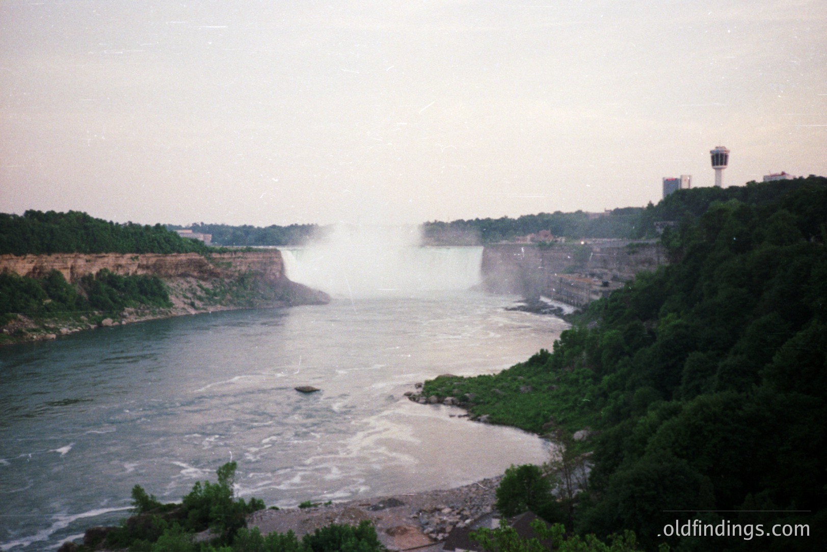 A wide, sweeping view captures Niagara Falls, partially obscured by mist and spray. Lush green vegetation clings to the rocky slopes flanking the powerful river flow. A tall observation tower is visible on the right. Likely captured on film, image has slight color cast.