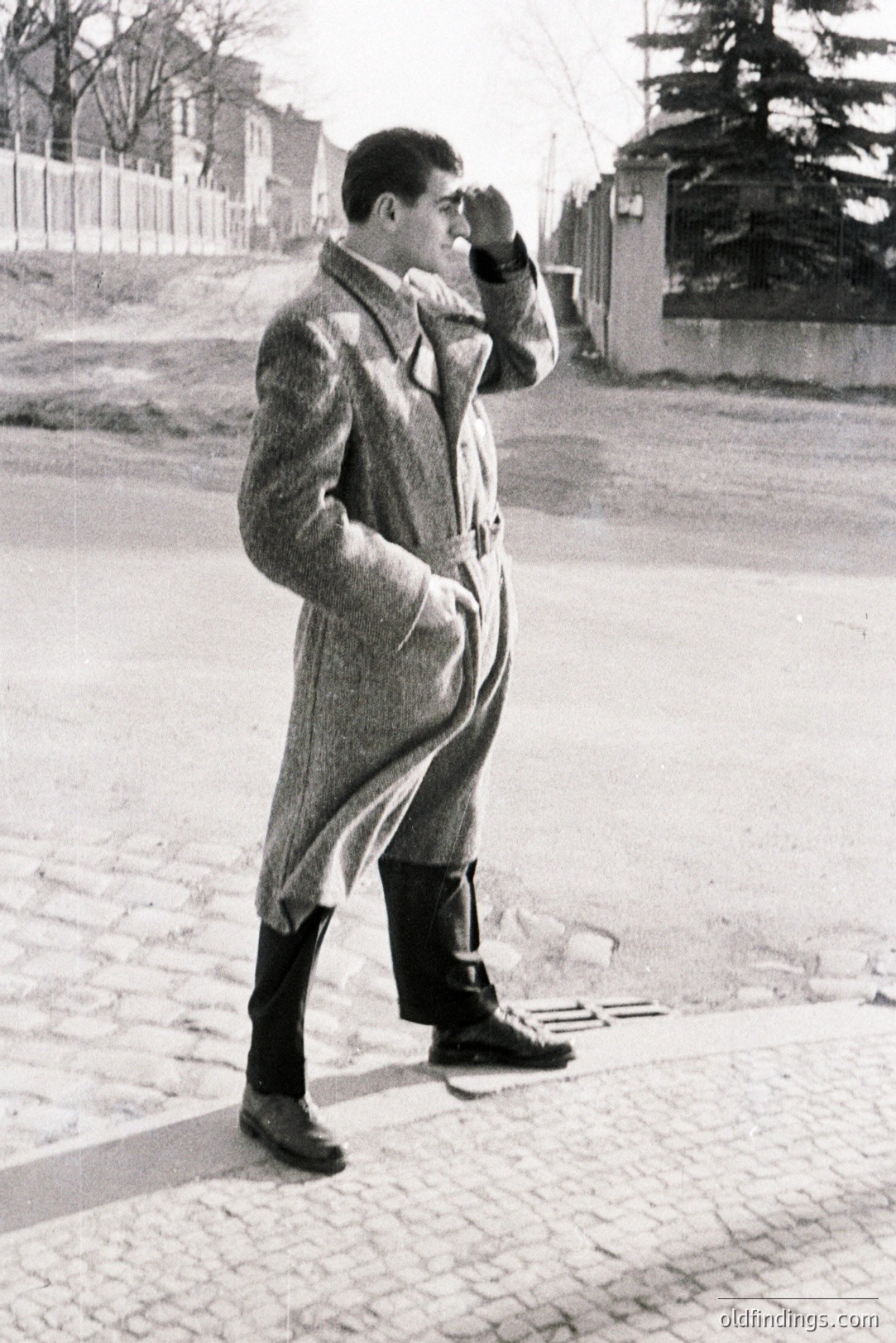 A man in a double-breasted coat and trousers stands on a cobblestone street, hand shielding his eyes. Classic 1960s style; likely European location with period architecture visible in the background. Potential stock image for fashion or historical design.
