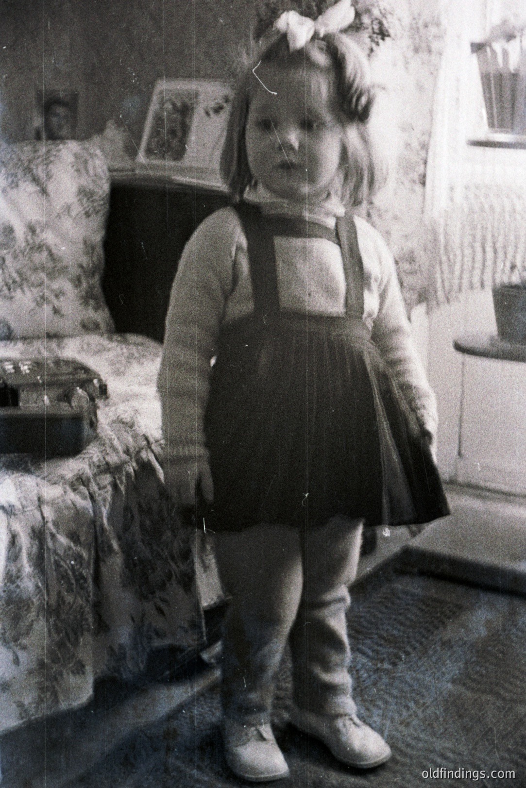 A young girl stands in a domestic interior, wearing a cable-knit sweater, dark overalls, and a ribboned headband. The room features floral wallpaper, a patterned armchair, and a glimpse of a window. Likely a family snapshot, 1960s-1970s style. Nostalgic, vintage aesthetic.