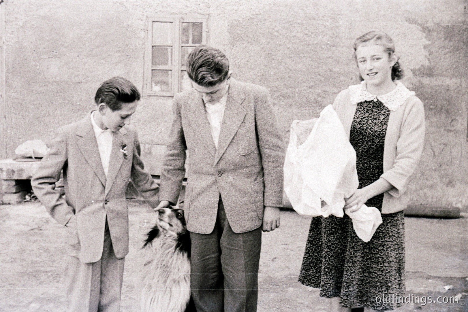 Three figures stand outside a building with a single window. A young boy in a suit, a young man in a suit, and a young woman in a dress holding a cloth. A dog stands between the two men. Likely a family portrait, mid-20th century. Possible Eastern European setting.
