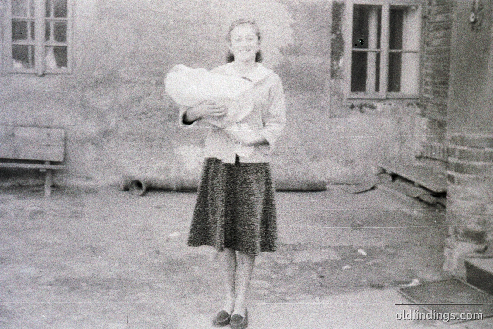 A woman stands in a courtyard, cradling a baby. She wears a patterned skirt and a light jacket. The architecture suggests a European setting, possibly post-war, judging by the visible damage and utilitarian design. Likely 1950s-1960s. Potentially valuable for historical research on family life.