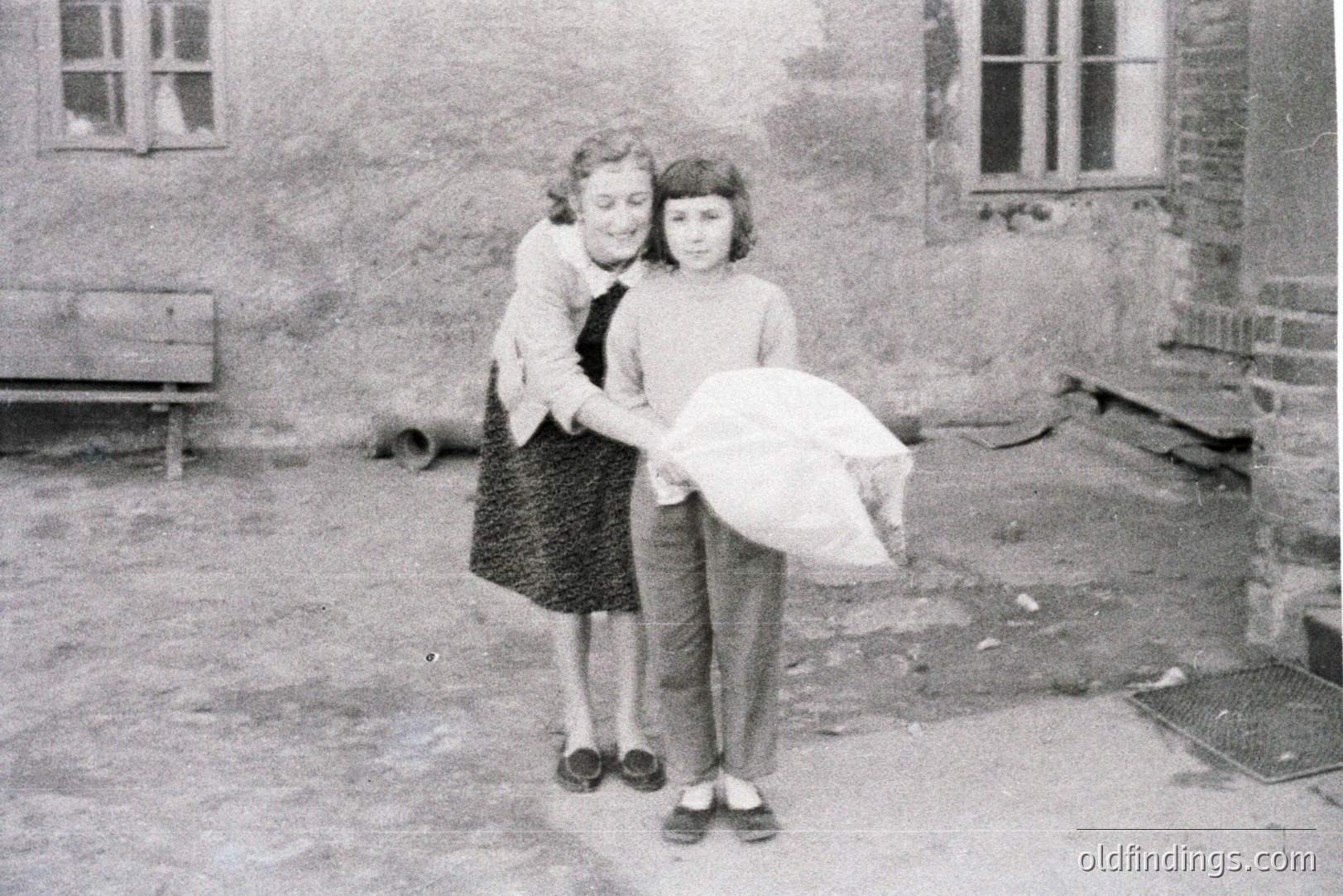 Two girls stand in a courtyard, one embracing the other who carries a large cloth bag. Brick buildings and a wooden bench form the backdrop. Likely mid-20th century, possibly post-war Europe. Simple, everyday scene with potential appeal for heritage or design use.