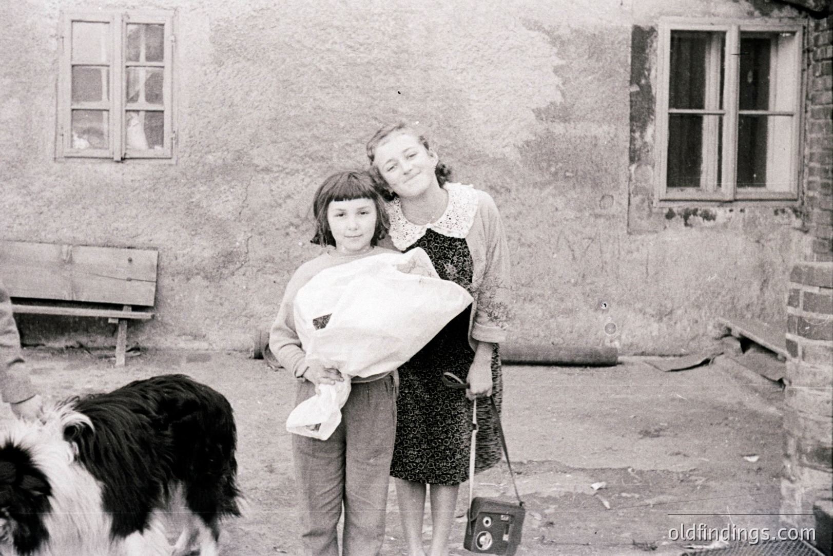 Two young girls stand outdoors, one cradling a wrapped bundle. A black and white border collie stands nearby. A weathered stone wall and simple architecture suggest a rural setting. The scene conveys a candid, domestic moment, likely 1950s. Appears to be a documentary or family snapshot.