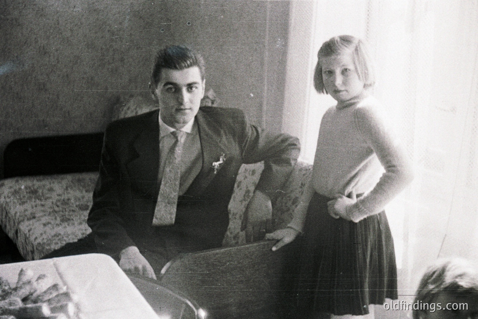 Man in suit & tie sits formally; a young girl stands beside him, hands on hips. Interior features patterned wallpaper & a bed. Likely a posed family portrait, c. 1950s-1960s. Simple elegance & restrained style evoke mid-century domesticity.