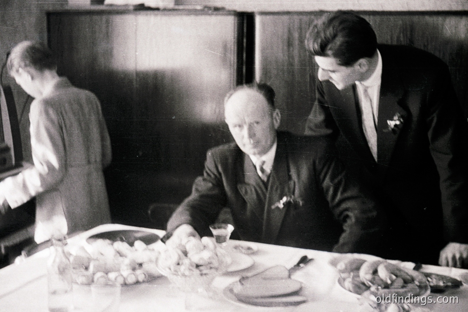 A formal dining scene: a seated man in a suit observes a lavish table laden with food and glassware. A younger man in a suit leans in. A figure in a dress is visible in the background. Likely a 1930s-40s family gathering, potentially documenting culinary traditions.