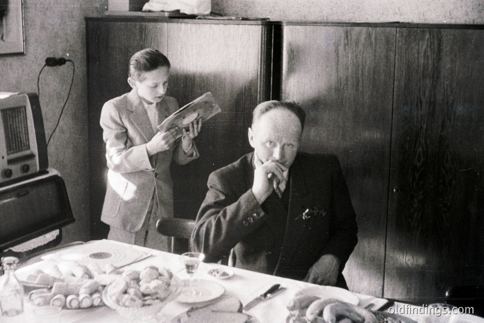 Man seated at a laden table, appearing contemplative. A young boy stands nearby, engrossed in a newspaper. Mid-century modern decor; likely a family gathering. The setting suggests a domestic interior, possibly late 1950s or early 1960s. A vintage radio is also visible.
