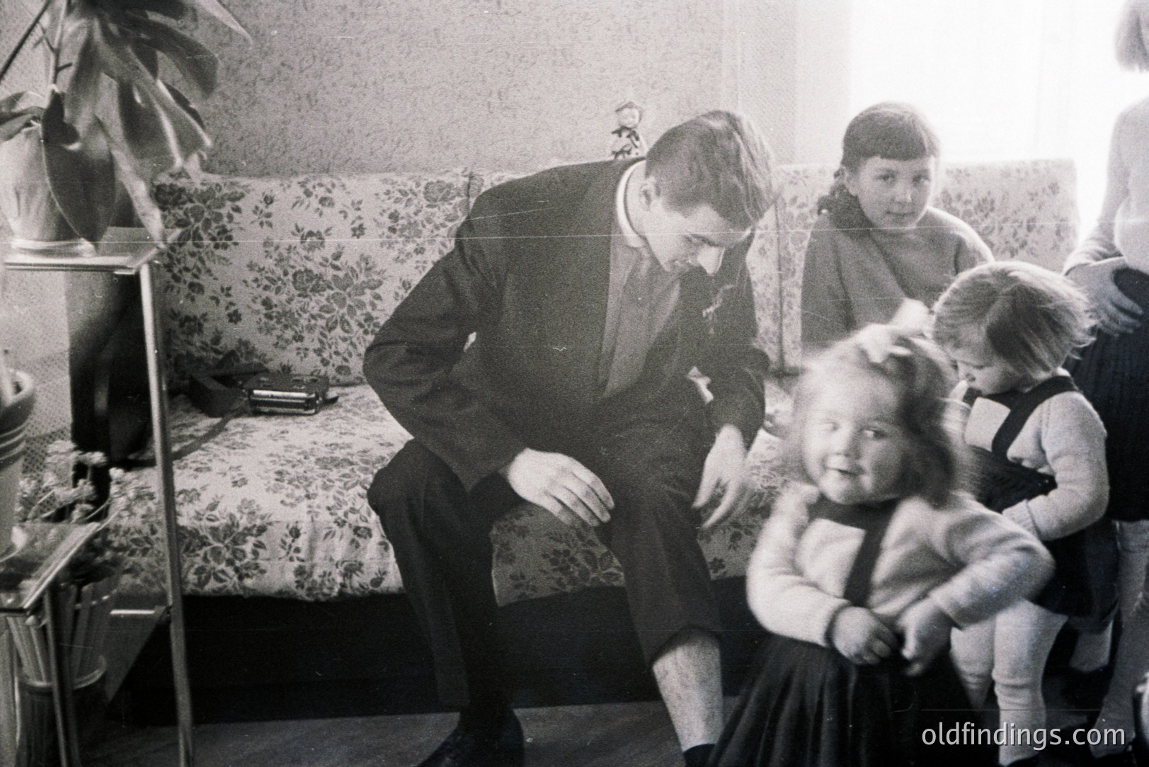 Man in a suit kneels to interact with two young girls. Floral patterned sofa, potted plant, and vintage furniture suggest a mid-century interior. Likely 1960s or 70s home life. Family portrait, potentially from Eastern Europe. Valuable for nostalgic design references.