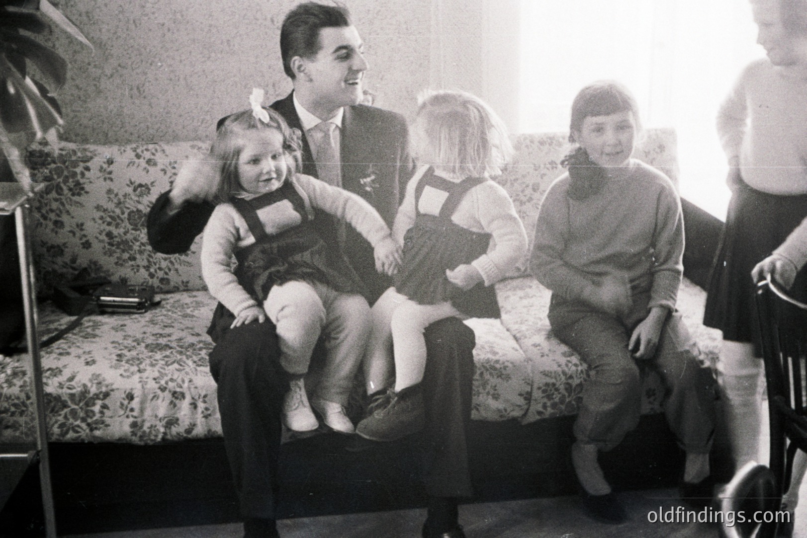Three young children sit on a man's lap on a floral patterned sofa. The children wear overalls and sweaters, with one playfully swatting at the man's face. Appears to be a domestic interior, potentially 1950s-1970s.