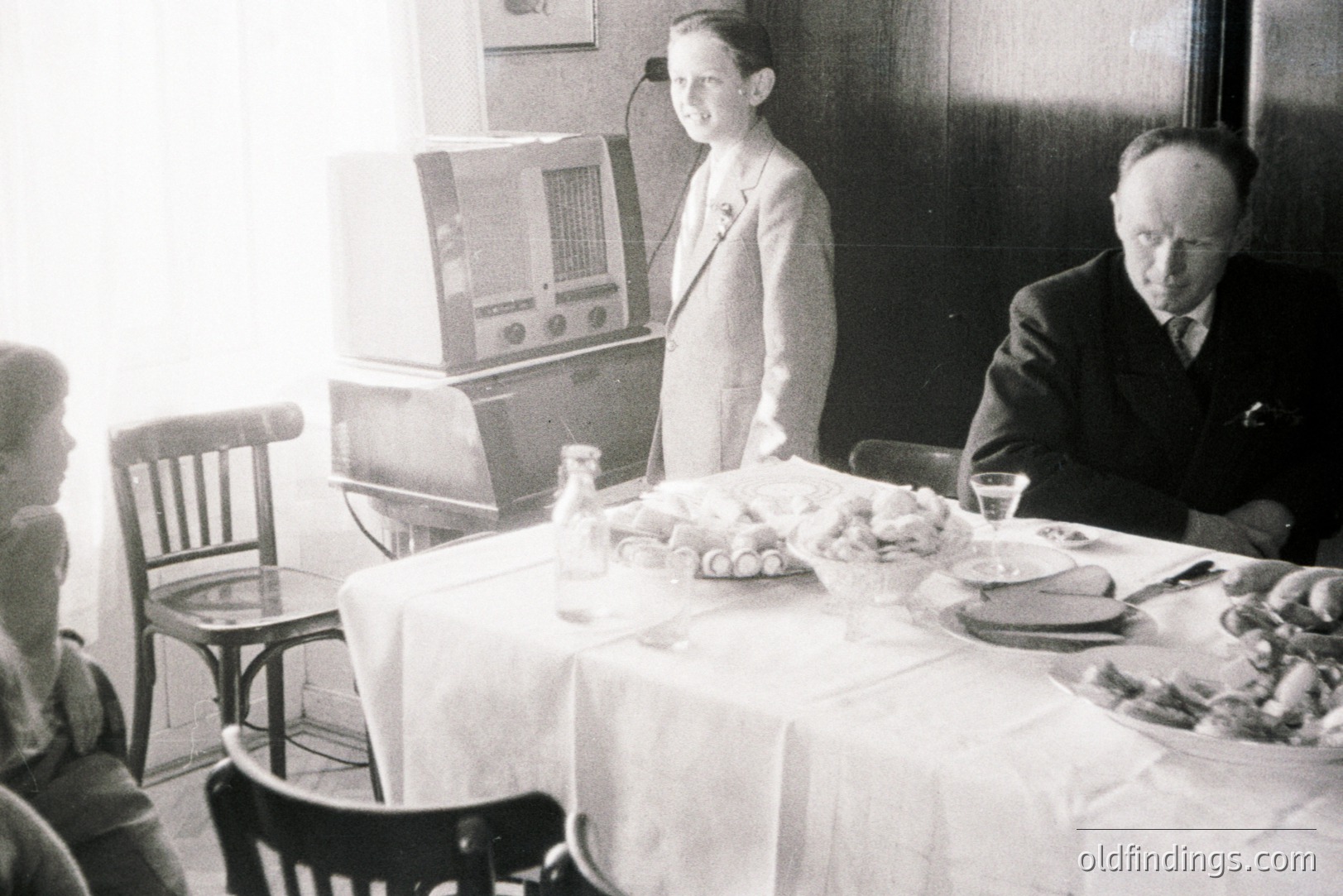 A family gathers around a table laden with food, featuring a vintage radio set prominently displayed. A young person stands near the radio, possibly presenting or demonstrating it. Likely taken in the 1930s, showcasing domestic life and early technology.