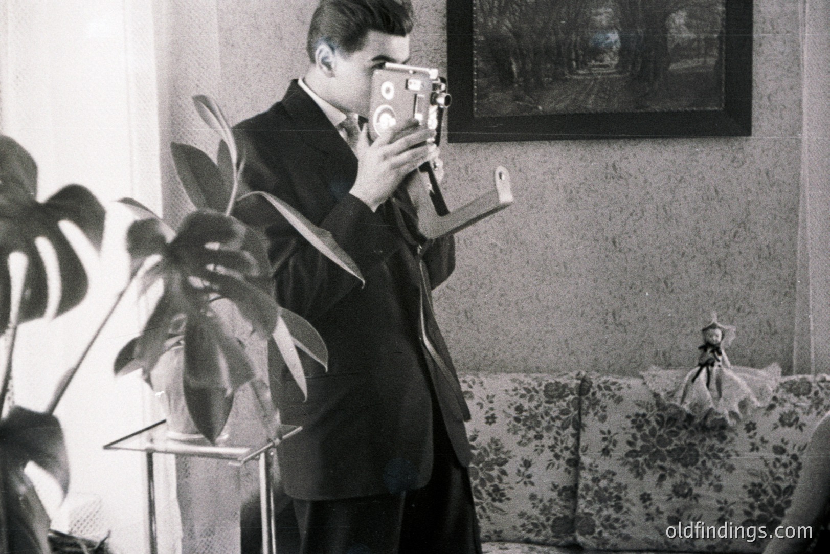 A sharply dressed man operates a vintage 8mm movie camera, likely filming within a domestic setting. The scene includes a potted plant, floral sofa, and framed artwork. The style suggests a 1950s or 60s home movie aesthetic. Could be stock footage reference.