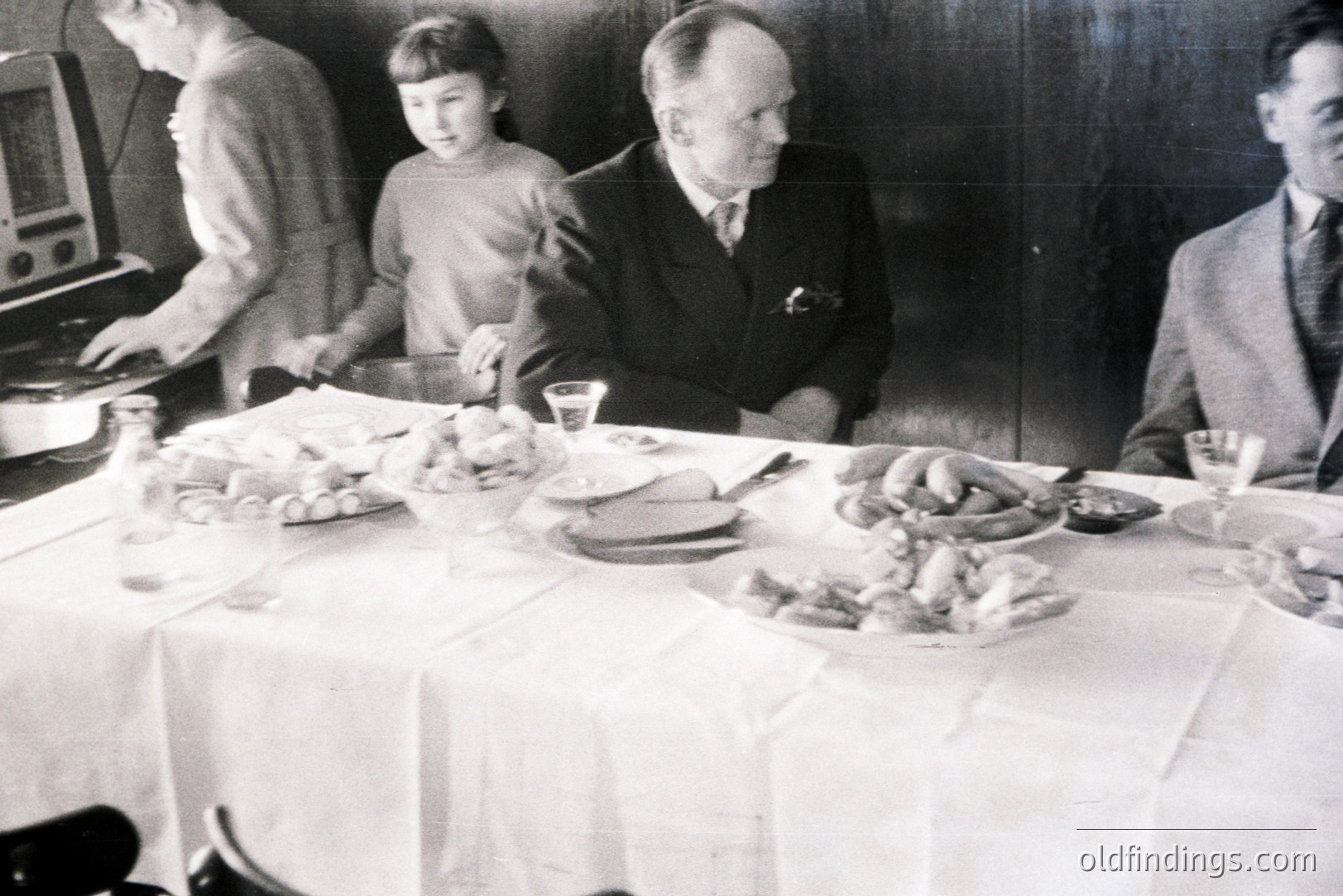 Family gathering around a long, draped table laden with food. A young girl stands near a vintage radio, while two formally dressed men look on. Likely a domestic scene, possibly a holiday or special occasion, 1940s-1950s.