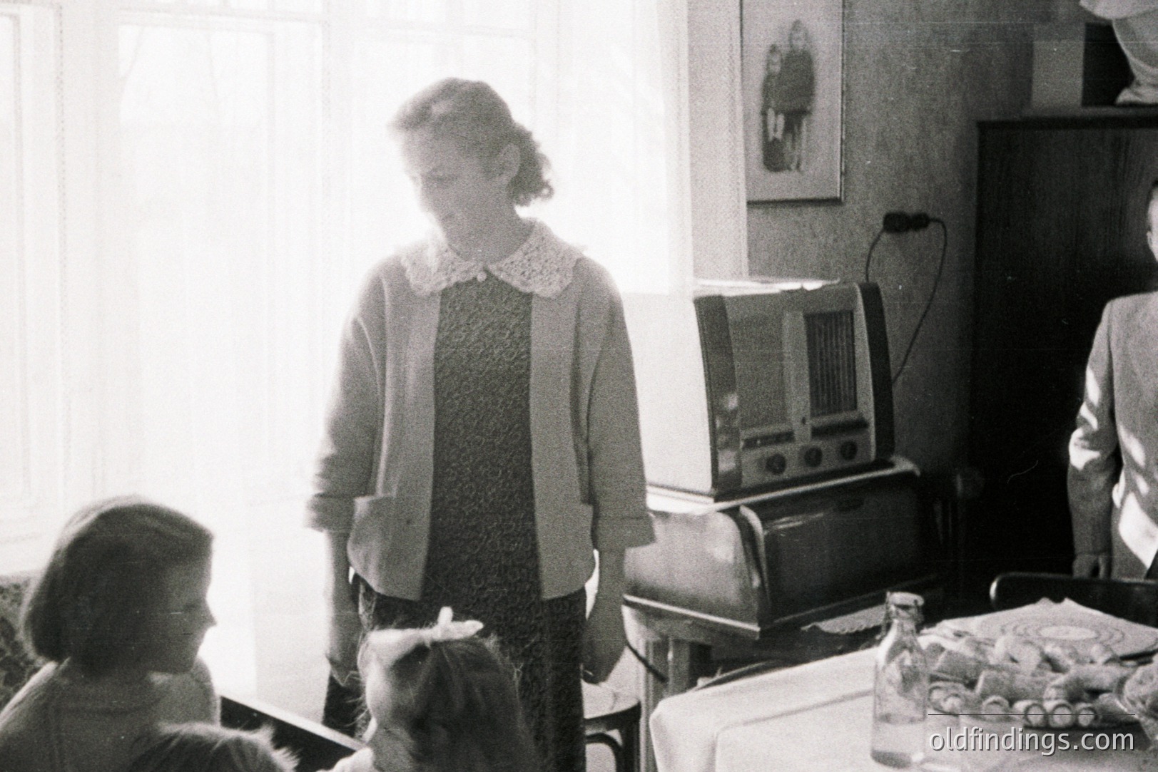 A seated young girl and a woman in a patterned dress and cardigan stand in a domestic interior. A large vintage radio dominates the scene, alongside a table set with glassware. Likely a mid-century family moment, captured in grayscale. Domesticity, radio technology, and childhood.
