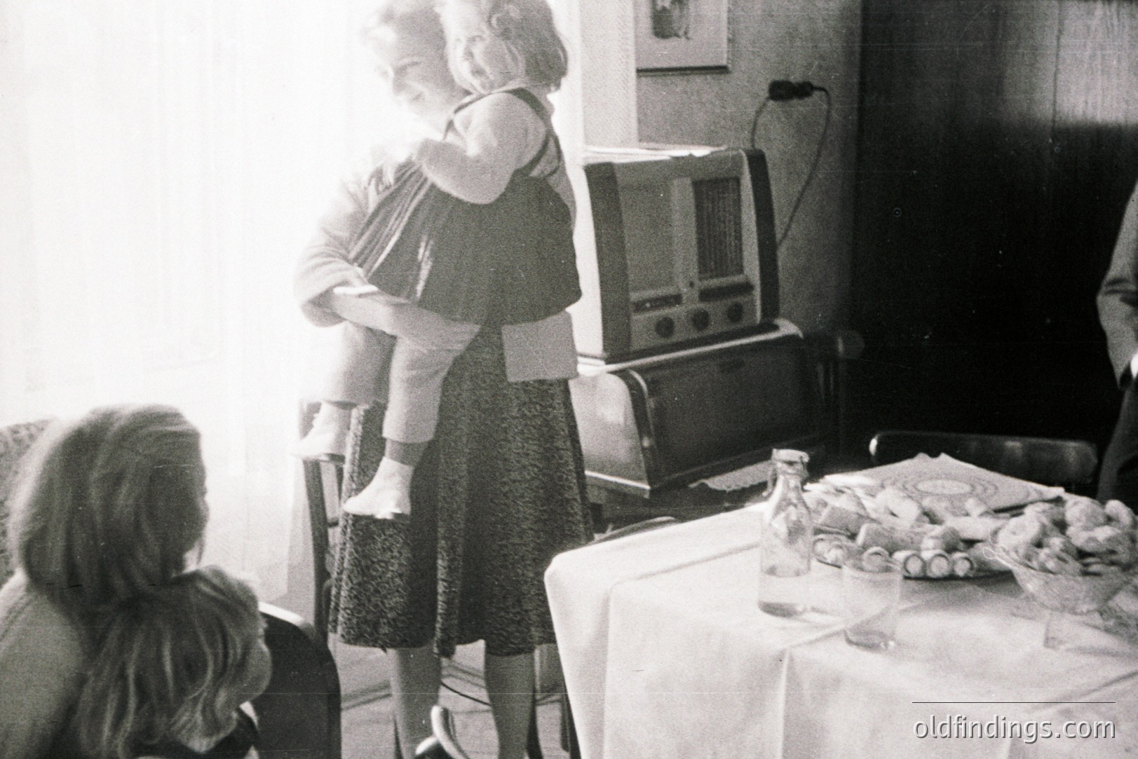 A woman in a patterned dress holds a small child on her hips, standing near a large, vintage radio console. A table with glassware and food is visible. A child’s back is partially visible in the foreground. Likely a family gathering, 1960s-1970s era. Domestic interior scene.
