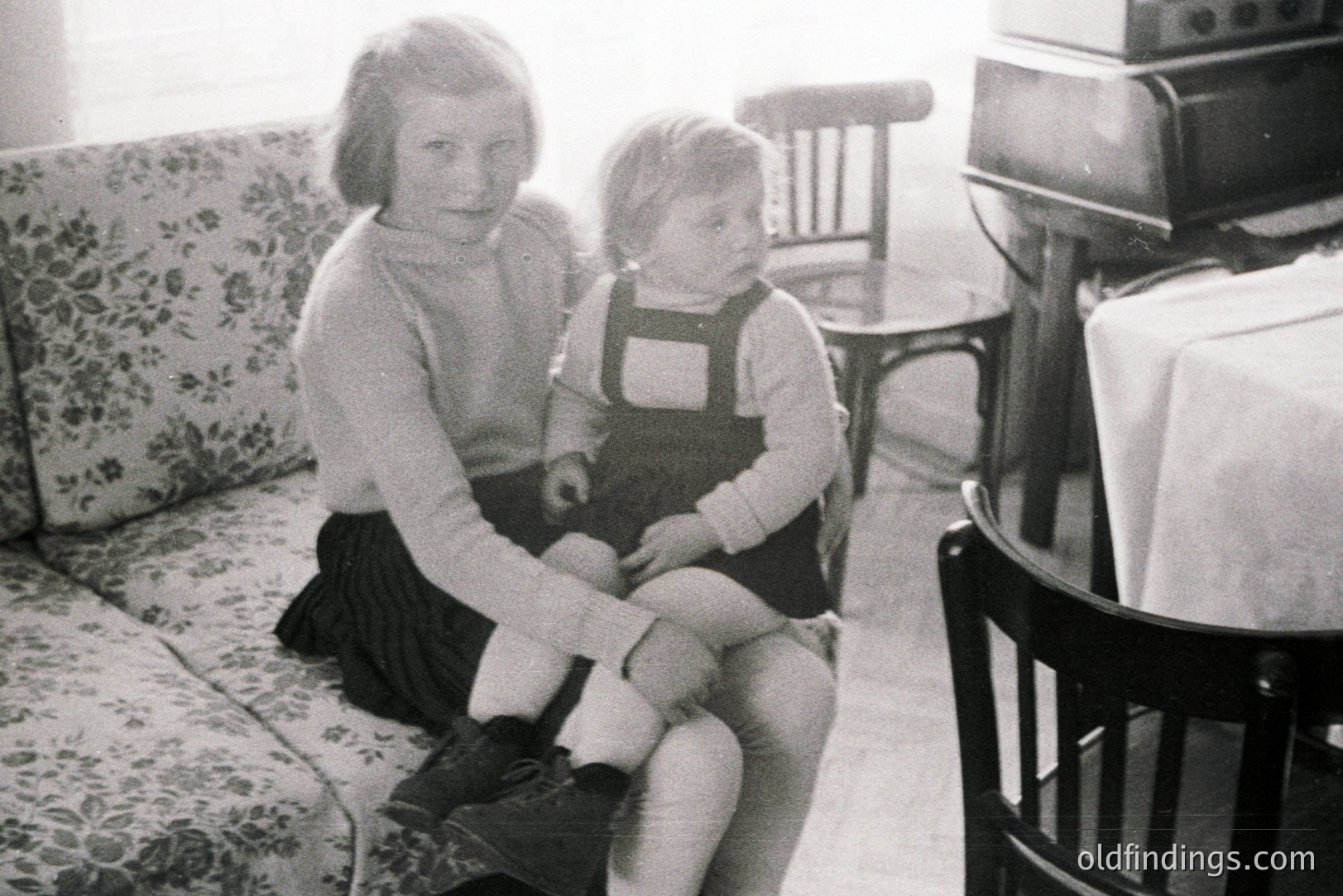 Two young girls, likely sisters, posed indoors. The older girl wears a pleated skirt & sweater; the younger sits on her lap in overalls & lace-up shoes. A floral-patterned sofa & a piano are visible. Likely a family snapshot, c. 1930s-1950s. Valuable for historical family studies and nostalgia marketing.