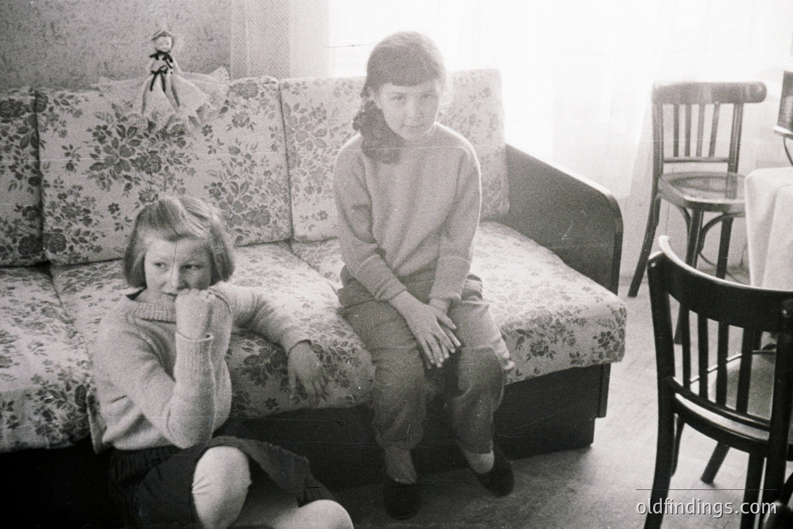 Two young girls in matching sweaters pose within a modestly furnished interior. One sits on a floral-patterned sofa, while the other is seated on the floor. A wooden chair and table are visible in the background. Likely a family snapshot, c. 1960s-1970s.