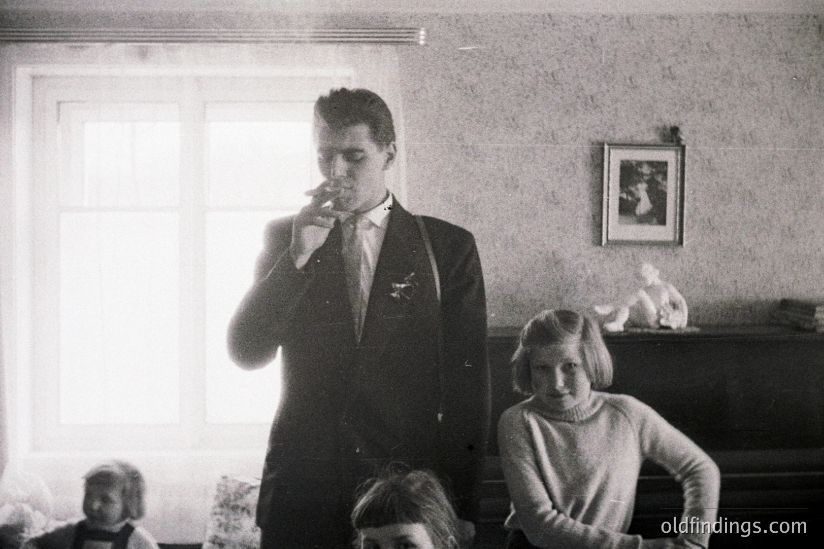 Formal portrait of a young man in a suit smoking, flanked by two girls seated near a piano. Wallpapered interior, likely a family home. The man's attire and the girls' hairstyles suggest a mid-20th century (1950s-1960s) setting. An ornate statue rests atop a piano.