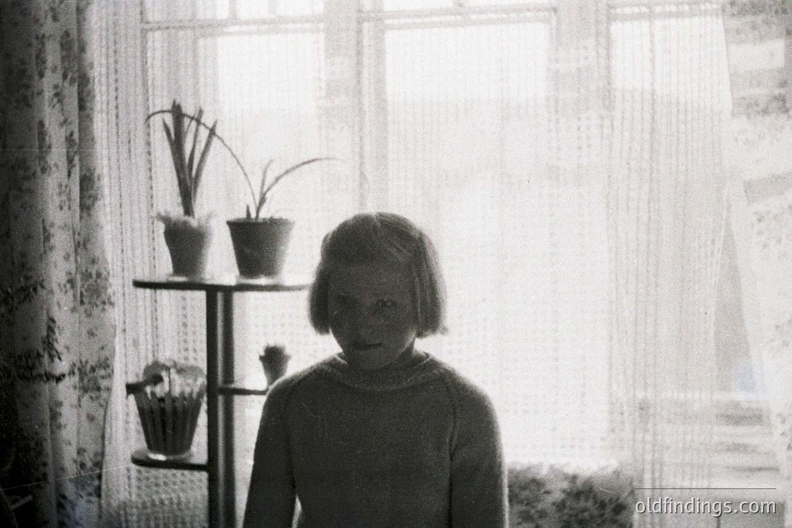 Young girl stands near a window, framed by sheer curtains & potted plants on a decorative shelving unit. Likely mid-20th century (1950s-1970s) domestic interior, possibly Scandinavia or Northern Europe. Simple, utilitarian style; evocative of post-war family life.