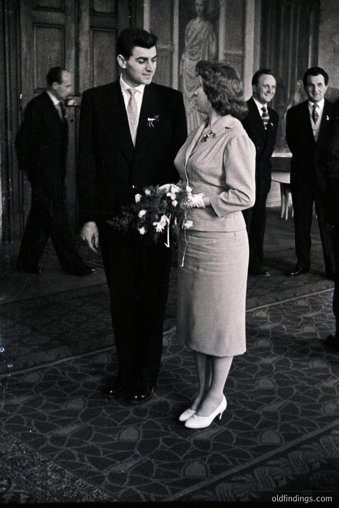 A formally attired couple stands close, likely at a wedding or official ceremony. The woman wears a light-colored suit and holds a bouquet of flowers, while the man wears a dark suit and tie. Several formally dressed men stand in the background. Likely 1960s-1970s.