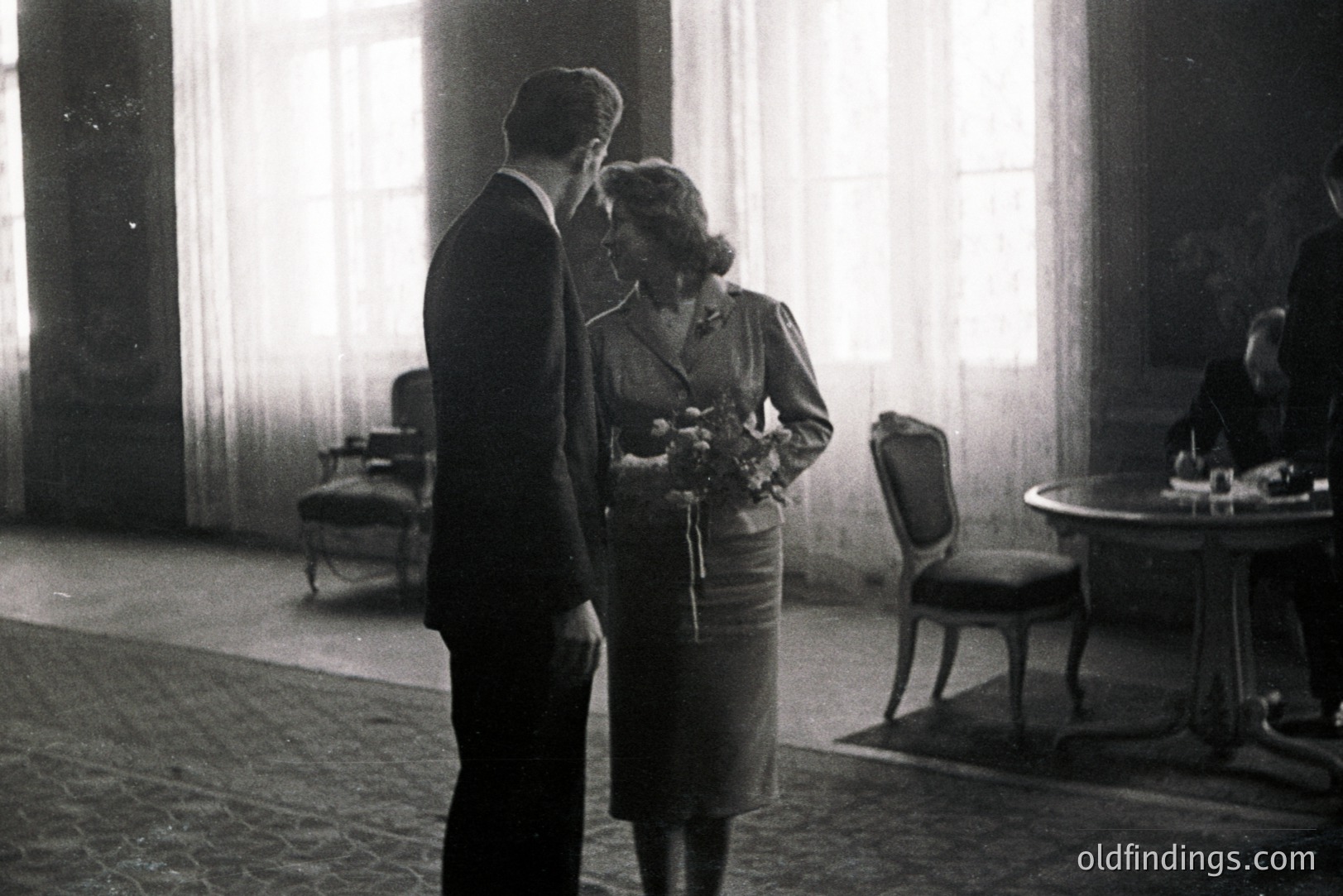 A formally dressed couple stands facing each other in a grand, light-filled room. The woman holds a bouquet of flowers. Visible through a window are snow-covered grounds. A seated figure is present in the background. Likely a 1950s or 60s portrait.